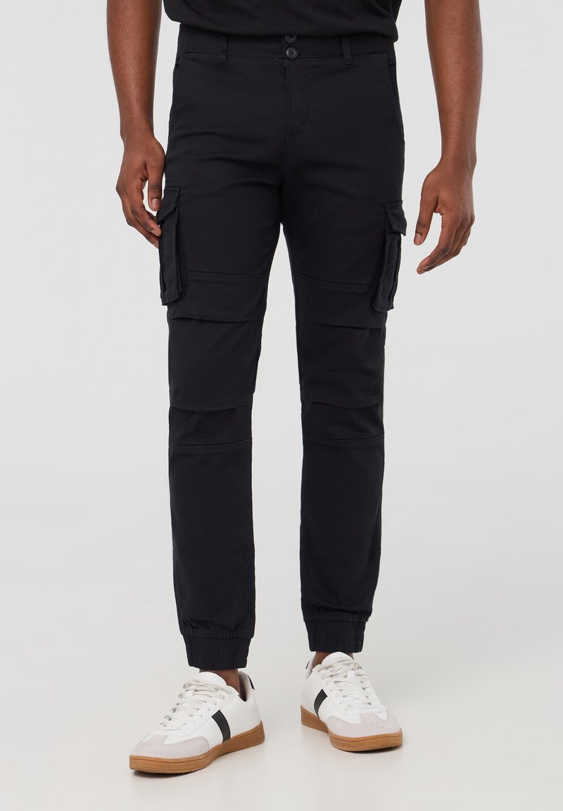 Czarne spodnie cargo mają wiele bocznych kieszeni, tapered fit, elastyczne mankiety i zapięcie na guzik w talii. Noszone z białymi trampkami.