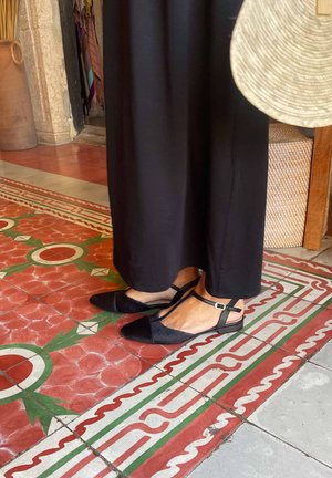 Kājām melnās smailās kurpēs ar potīšu siksniņām, stāvot uz sarkanas, zaļas un baltas rakstainas flīžu grīdas blakus pinumam grozam un salmu cepurei.