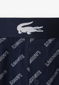 Navy blue shorts featuring a waistband with white Lacoste logo and repeated text pattern, made of lightweight fabric with a soft texture.