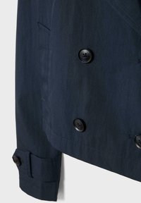 Manche d'une veste bleu marine foncé et bas de devant avec boutons noirs et petite poche fendue près de la couture latérale.