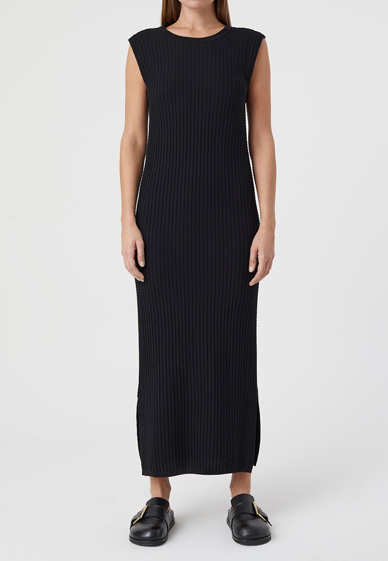 Schwarz geripptes Maxikleid mit Rundhalsausschnitt und kurzen Ärmeln. Verfügt über seitliche Schlitze für mehr Bewegungsfreiheit. Getragen mit schwarzen Slippern.