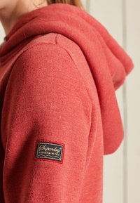 Sudadera roja con un tejido suave, una capucha grande y un parche de logo negro de Square Superdry en la manga izquierda.