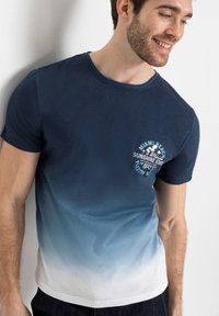 Maglietta a maniche corte con un gradiente dal blu navy al bianco, caratterizzata da un design grafico azzurro sul petto. Tessuto morbido.
