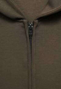 Smeđa zip-up dukserica od teksturiranog materijala, s punim crnim patentnim zatvaračem i kapuljačom za dodatnu zaštitu.