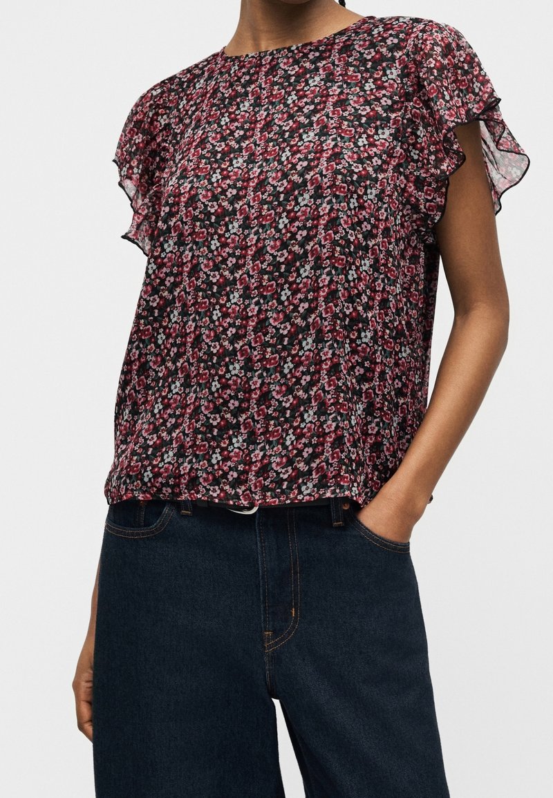 Femme portant un chemisier court à manches courtes transparent à fleurs avec des manches volantées et un jean taille haute bleu foncé, main dans la poche.