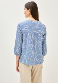 Blouse à motifs bleus avec des manches trois-quarts, présentant un léger motif de feuilles et un tissu texturé, associée à un pantalon crème.
