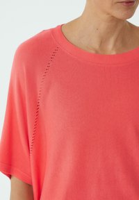 Koronkowa bluzka w kolorze koralowym z okrągłym dekoltem, luźnym krojem i krótkimi rękawami. Posiada dekoracyjne perforacje wzdłuż szwu na ramieniu.