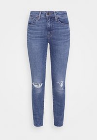 Kapeat farkut keskisinisessä denimissä, joissa on korkea vyötärö, lievää kulutusta ja kulutettu polviosuus. Klassinen viiden taskun muotoilu.