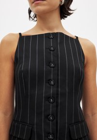 Ženska v črnem brezrokavniku s podolžnimi črtami, ki ima sedem gumbov spredaj in žepke s klapnico na pasu.