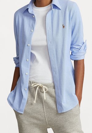 Person iført en lyseblå button-up skjorte over en hvid t-shirt med grå snøre bukser, hænderne i lommerne, skjorteærmerne opkrogede.