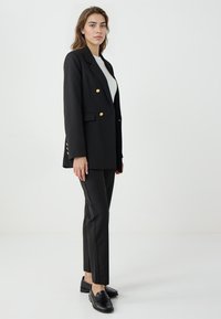 Zwarte dubbel geknoopte blazer met gouden knopen, revers en zijzakken, gecombineerd met slanke zwarte broeken en zwarte instappers, staand tegen een lichte achtergrond.