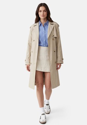 Vrouw in beige trenchcoat over een blauw gestreept hemd en een beige rok, lopend op witte sneakers met zwarte strepen, tegen een witte achtergrond.