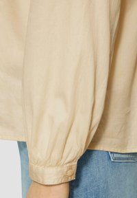 Beige katoenen blouse met opgestoken lange mouwen, gathered manchetten en een knoopsluiting. Gekozen over lichtblauwe denim. Soepele textuur.