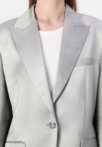 Helehall blazer sujuva tekstuuriga, kaldus kraega, ühe nööbiga kinnitamine ja eesmine tasku, kantud ribaga valge särgi üle.