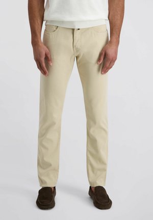 Mann trägt beige schlanke Hosen und dunkelbraune Slipper, steht vor einem einfarbigen hellen Hintergrund.