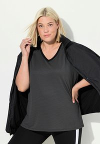 Černý sportovní top s V-čelním výstřihem a strukturovaným vzorem, nošený pod černou bundou. Modelka má rovné blond vlasy a pózuje s jednou rukou na bocích.