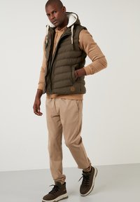 Olive green puffer vest with a zippered front, cream hood lining, tan sweatshirt underneath, khaki pants, and brown lace-up boots.