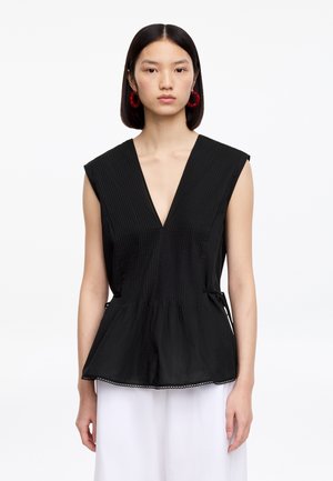 Jeune femme aux cheveux noirs raides portant des boucles d'oreilles rouges en forme d'anneau, un haut noir sans manches à col en V avec des plis, et un pantalon blanc, debout devant un fond blanc.