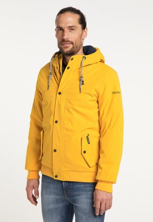 Mann mit Bart, der eine gelbe Kapuzenjacke mit Kordelzug und blaue Jeans trägt und vor einem schlichten Hintergrund steht.