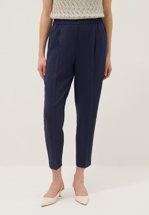 Vrouw die marineblauwe taps toelopende broek met plooien draagt, een beige mouwloze gebreide top en crèmekleurige puntige kitten heels.