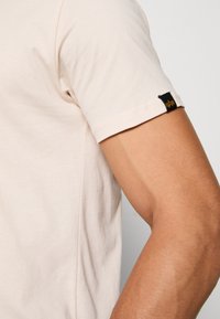 Beige t-shirt med korte ærmer; blødt bomuldsstof; sort mærke med gyldent logo på ærmet; glat tekstur; tætsiddende design.