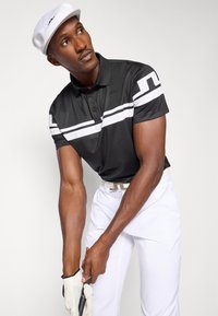 Μαύρο πόλο μπλουζάκι με λευκές ρίγες, κοντό μανίκι και γιακά. Συνδυασμένο με λευκά παντελόνια και καπέλο ανοιχτού χρώματος. Κρατώντας μια ράβδο γκολφ.