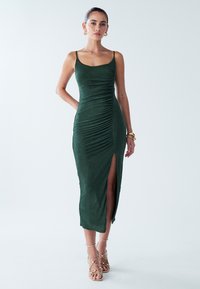 Eng anliegendes grünes Kleid aus strukturiertem Stoff mit gerafften Details, dünnen Trägern und einem Schlitz bis zur Mitte des Oberschenkels. Hochwertige Schuhe sichtbar.