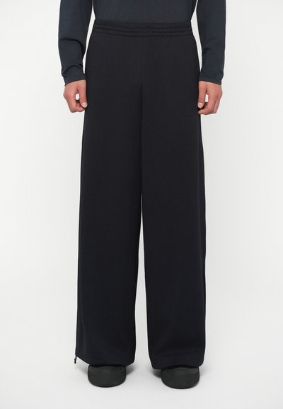 Schwarze, weit geschnittene Hosen aus einem strukturierten Stoff mit einem elastischen Bund. Mit Reißverschlüssen an den Knöcheln für eine verstellbare Passform.
