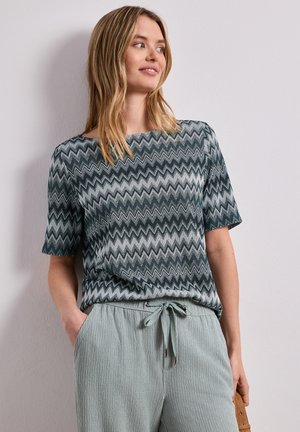 Frau trägt ein kurzärmeliges, zickzackgemustertes Oberteil und helle, strukturierte Hose mit Kordelzug, steht entspannt mit einer Hand in der Tasche.