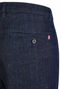 Donkerblauwe denim broek met een achterzak met knoopsluiting en een klein rood, wit en blauw stoffen label dicht bij de rand van de zak.