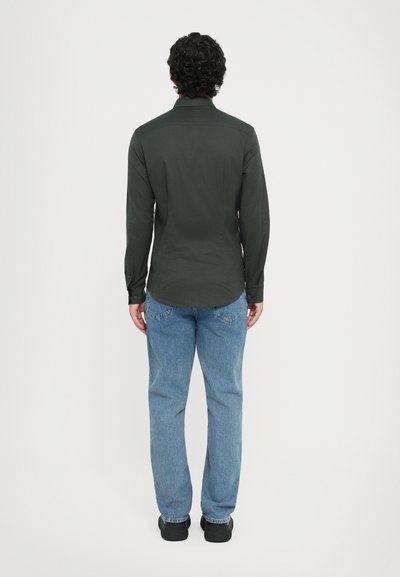 Cămașă verde închis cu mânecă lungă, cu guler, cu o textură fină, purtată cu blugi denim albaștri deschiși și pantofi negri.