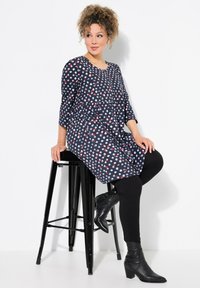 Túnica azul oscura con un patrón en blanco, rojo y azul claro, de mangas tres cuartos, corte relajado, combinada con leggings negros y botas de tobillo.