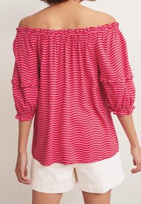 Rød off-shoulder bluse med hvide bølgede striber, pufærmer med flæsekant, parret med hvide shorts. Blødt stof, afslappet pasform.