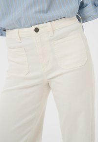 Primo piano di pantaloni bianchi a vita alta con tasche frontali, passanti per cintura e chiusura con bottone, indossati con una camicia a righe blu infilata dentro.