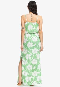 Zelené květinové maxi šaty s bílými vzory hibiskusu, tenkými ramínky a bočním rozparkem. Vyrobeno z lehkého, prodyšného materiálu.