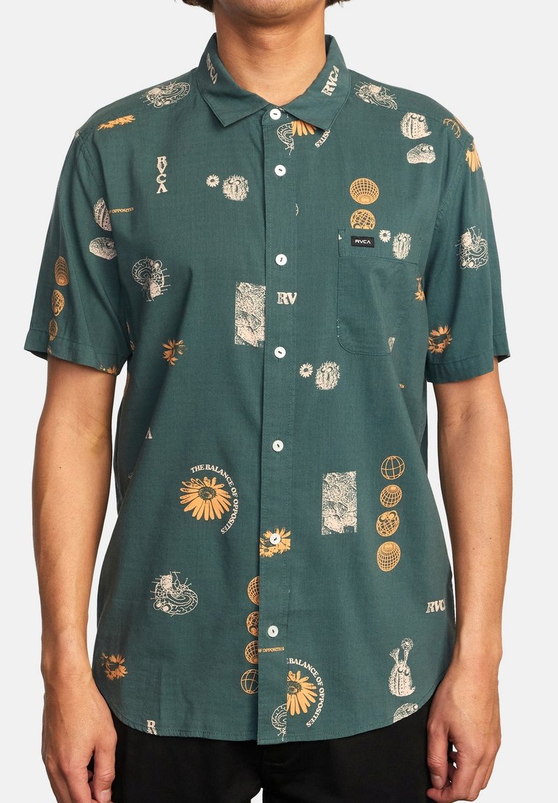 Homme portant une chemise verte à manches courtes avec des motifs abstraits et floraux orange et blancs et une poche poitrine.