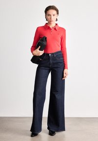 Cămașă polo roșie cu mânecă lungă, cu guler și detalii cu nasturi, asortată cu pantaloni din denim evazați de culoare bleu închis și ținând o geantă de mână neagră.