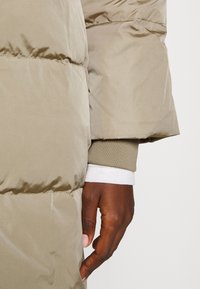 Kaki pufferjakke med quiltet struktur, der har en tætsiddende manschetærme og lagdelt stof ved håndleddet, som viser et kontrasterende lys lag under.