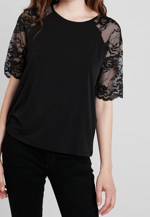 Vrouw die een zwarte top draagt met doorschijnende kanten korte mouwen met bloemenpatroon en een zwarte broek, staande tegen een effen grijze achtergrond.