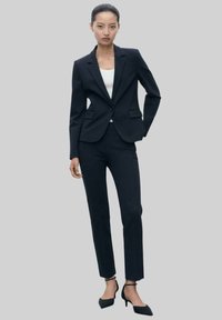Navy räätälöity bleiseri, jossa on lovetut kaulukset, yhdistettynä yhteensopiviin slim-fit-housuihin. Mallilla on päällään valkoinen paita ja terävät mustat korot.