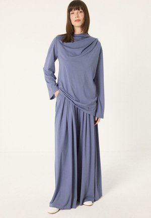 Femme debout portant un haut bleu à manches longues ample et un pantalon large bleu avec des chaussures blanches sur fond uni.