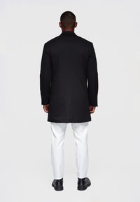 Zwarte jas met een scherpe snit, gecombineerd met witte broek. De jas heeft een gladde textuur en een klassieke kraag, met lange mouwen en knopen.