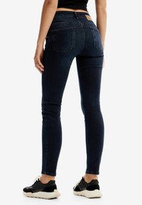 Jeans ajustados de denim azul oscuro, con una cintura de altura media, bolsillos traseros y una sutil textura desgastada. Combinados con zapatillas deportivas negras.