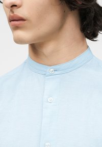 Κοντινό πλάνο ενός ατόμου που φοράει ένα ανοιχτό γαλάζιο πουκάμισο με γιακά μπάντας και λευκά κουμπιά, εμφανίζοντας τη γραμμή του σαγονιού και την περιοχή του λαιμού.