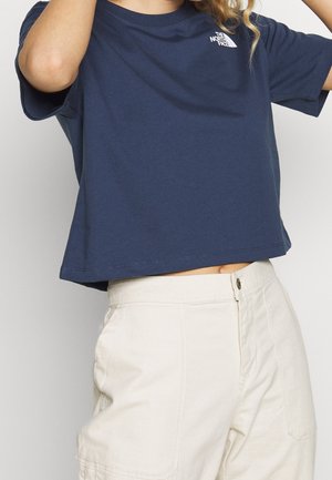 Persoon draagt een donkerblauw cropped North Face t-shirt met beige hoog taille broeken, armen gedeeltelijk opgeheven, tegen een effen achtergrond.