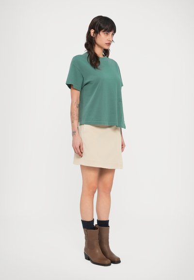 Πράσινο κοντομάνικο t-shirt με άνετη εφαρμογή, συνδυασμένο με μπεζ φούστα και καφέ μποτάκια αστραγάλου. Ορατές υφασμάτινες κάλτσες πάνω από τα μποτάκια.