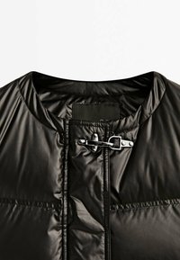 Veste matelassée noire brillante avec un col rond et une fermeture à crochet en métal argenté en haut devant.