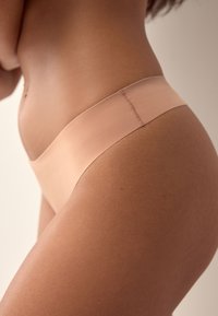 Nærbillede af en kvinde iført sømløst undertøj i nøgenfarve, der viser hendes hofte og nedre talje mod en ensfarvet baggrund.