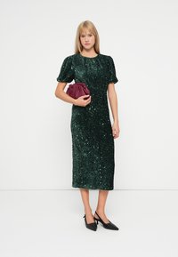 Vestido midi em verde esmeralda com lantejoulas e mangas volumosas, combinado com uma carteira bordô. Mulher a usar saltos pretos pontiagudos.