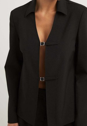Black long-sleeve shirt with collar, fastened by two small silver buckles at the chest, worn open to reveal skin underneath.